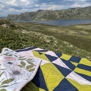 The 279 Quilt Pattern (Download) by Millie & Bun Designs displayed outdoors, showcasing geometric and floral quilt patterns with a mountainous lake backdrop.