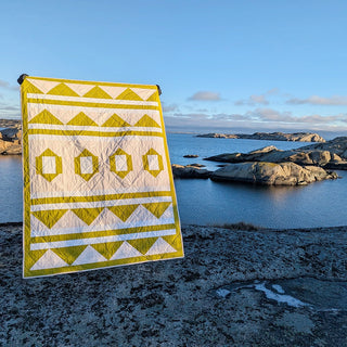 Quilt with geometric yellow and white pattern hanging outdoors near rocky coastline under clear blue sky, showcasing The 2995 Quilt Pattern by Millie & Bun Designs.