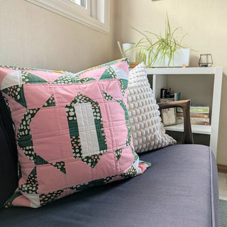 A green and pink decorative quilted pillow featuring a geometric pattern made with The 2995 Quilt Quilt Pattern (Download) by Millie & Bun Designs, displayed on a gray sofa.