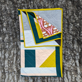 Folded modern quilt with geometric shapes and contrasting colors, showcasing a floral patterned corner, made using The 3895 Quilt Pattern (Download) by Millie & Bun Designs.