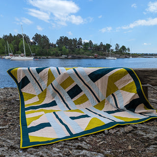 The 3895 Quilt Pattern (Download) by Millie & Bun Designs displayed outdoors by a lake, featuring a modern geometric design in green, yellow, and white tones.