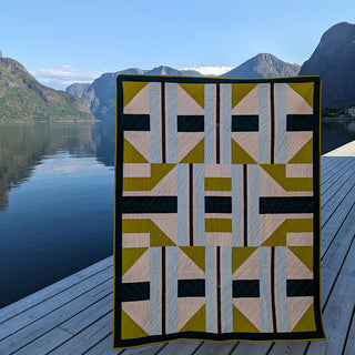 The 3895 Quilt Pattern (Download) by Millie & Bun Designs features a modern geometric design with olive green, black, beige, and blue tones displayed against a scenic mountain lake backdrop.