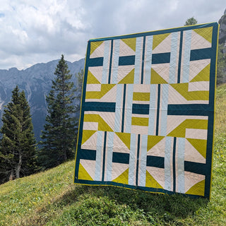 A modern geometric quilt in soft blue, green, and cream tones displayed outdoors against a mountain backdrop, showcasing The 3895 Quilt Pattern (Download) by Millie & Bun Designs.