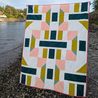 A geometric quilt in shades of teal, mustard, pink, and white, displayed outdoors near water, showcasing The 5720 Quilt Pattern (Download) by Millie & Bun Designs.