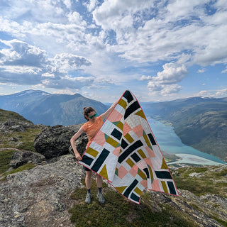 Woman holding a geometric quilt made with The 5720 Quilt Pattern (Download) by Millie & Bun Designs, standing on a mountain with scenic clouds and lake views.