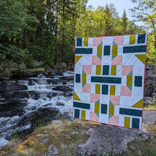 A vibrant modern quilt featuring geometric shapes in pink, green, white, and mustard, displayed outdoors by a woodland stream, showcasing The 5720 Quilt Pattern (Download) by Millie & Bun Designs.
