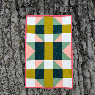 Colorful geometric quilt with pink, green, yellow, white, and dark teal shapes showcasing The 5720 Quilt Pattern (Download) by Millie & Bun Designs hanging on tree bark.