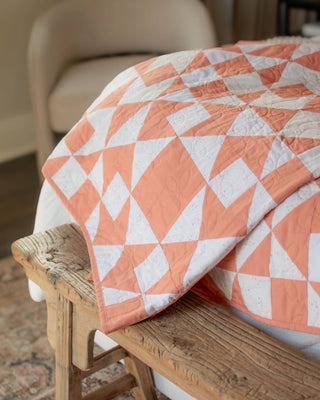 A peach and white geometric quilt draped over a wooden bench in a cozy room, showcasing the Afterglow 2.0 Quilt Pattern (Download) by Sewspicious.