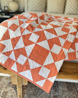 Orange and white geometric quilt with intricate stitching displayed on a wooden table, showcasing Sewspicious craftsmanship using Afterglow 2.0 Quilt Pattern (Download).
