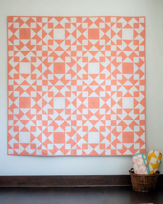 A peach and white geometric quilt hanging on a wall with a basket of colorful rolled quilts nearby, featuring the Afterglow 2.0 Quilt Pattern (Download) by Sewspicious.