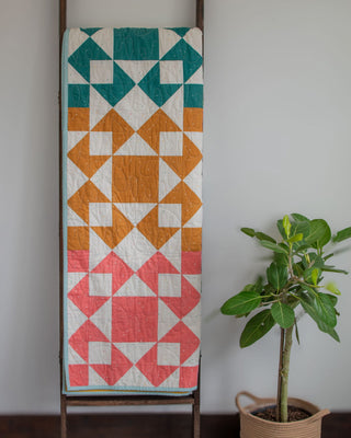 Colorful geometric quilt hanging on a ladder next to a potted plant, showcasing the Afterglow 2.0 Quilt Pattern (Download) by Sewspicious.