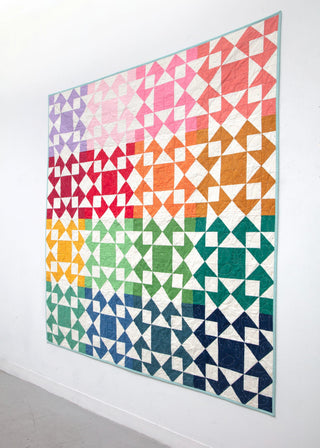 Colorful quilt hanging on a white wall with geometric blocks in a rainbow gradient, showcasing the Afterglow 2.0 Quilt Pattern (Download) by Sewspicious.