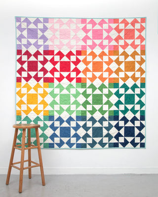 Colorful geometric quilt with gradient hues arranged in star and triangle blocks, displayed on a white wall next to a wooden stool, showcasing the Afterglow 2.0 Quilt Pattern (Download) by Sewspicious.