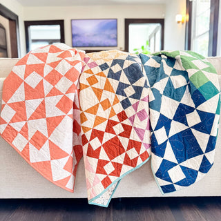 Three colorful geometric quilts draped over a couch showcasing bold triangular and diamond shapes in orange, blue, green, and red tones, made using the Afterglow 2.0 Quilt Pattern (Download) by Sewspicious.