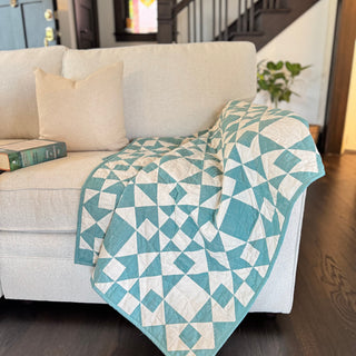 A cozy cream and teal geometric quilt draped over a beige couch with a matching pillow, showcasing the Afterglow Quilt Pattern (Download) by Sewspicious.