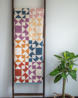 Afterglow Quilt Pattern (Download) by Sewspicious displayed on a wooden ladder, featuring vibrant geometric blocks in shades of purple, orange, blue, and cream.