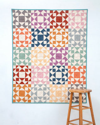 Colorful geometric patchwork quilt hanging on a wall next to a wooden stool, showcasing the Afterglow Quilt Pattern (Download) by Sewspicious in a modern, scrappy style.
