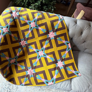 Colorful geometric quilt draped over a gray tufted couch, showcasing a detailed pattern with yellow, brown, pink, and blue hues, created using the Alcove FPP Quilt Pattern (Download) by Julia Wachs Designs.