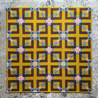 Colorful quilt with geometric star and cross patterns in mustard, brown, pink, and teal, displayed on a textured floor, showcasing the Alcove FPP Quilt Pattern (Download) by Julia Wachs Designs.