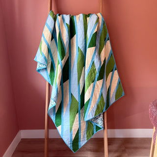 Quilt with modern geometric design in blue, green, and white tones displayed on wooden ladder in cozy room, showcasing Alderbrook Quilt Pattern (Download) by Julia Wachs Designs.