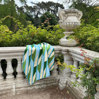 A colorful quilt with green, blue, and cream geometric patterns draped over a stone railing in a garden, showcasing the Alderbrook Quilt Pattern (Download) by Julia Wachs Designs.