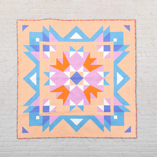 Colorful geometric quilt with orange, pink, blue, and white shapes arranged in a star pattern on a peach background, showcasing the Aria Quilt Pattern (Download) by Toad & Sew.