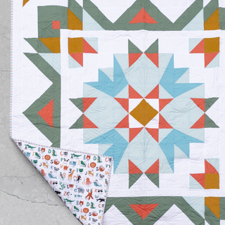 Close-up of a colorful geometric quilt with green, blue, orange, and white blocks, featuring a patterned fabric backing, showcasing the Aria Quilt Pattern (Download) by Toad & Sew.