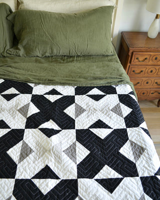 Modern black, white, and gray geometric quilt displayed on a bed with olive green bedding and wooden nightstand, showcasing the Bedroom Quilt Pattern (Download) by Sewn.