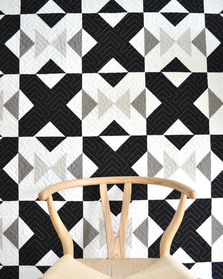 Black, white, and gray geometric quilt pattern displayed behind a light wood chair, showcasing the Bedroom Quilt Pattern (Download) by Sewn with a clean, modern design.