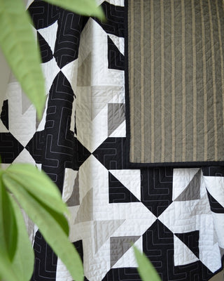 Close-up of a modern black, white, and gray geometric quilt draped with a striped gray fabric, showcasing the Bedroom Quilt Pattern (Download) by Sewn with visible quilting details.
