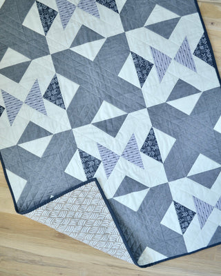 Bedroom Quilt Pattern by Sewn grays colorway: grey-toned flying geese block quilt with geometric X shapes, photographed from a corner angle on a wood floor.