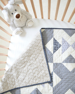 Soft grey and white baby crib quilt with geometric patterns, styled with a plush stuffed dog, showcasing the Bedroom Quilt Pattern (Download) by Sewn.