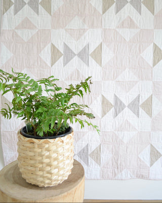 Soft neutral quilt with geometric pattern displayed behind green fern in woven basket, showcasing Bedroom Quilt Pattern (Download) by Sewn.
