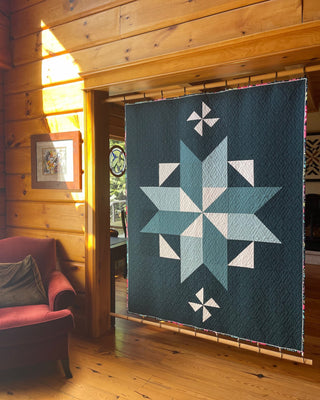 Quilt hanging inside a cozy wooden room featuring a geometric star design in blues and white, showcasing the Black Balsam Quilt Pattern (Download) by Hampton Quilt Studio.