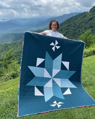 Black Balsam Quilt Pattern by Hampton Quilt Studio cover: person holding a teal and navy large star-block quilt in front of a mountain view.