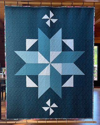 Quilt hanging on a wooden frame featuring a large eight-point star and smaller pinwheel designs in shades of blue and white, showcasing the Black Balsam Quilt Pattern (Download) by Hampton Quilt Studio.