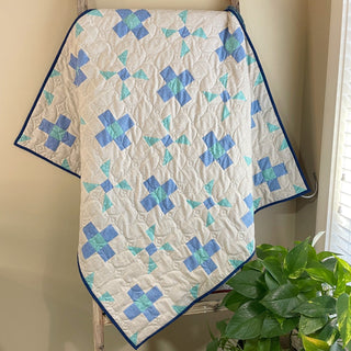 Light quilt with blue and teal geometric blocks hanging on a ladder, showcasing the Breezy Shores Quilt Pattern (Download) by Shadow Valley Quilts in a cozy room setting.