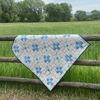Breezy Shores Quilt Pattern (Download) by Shadow Valley Quilts shows a fresh blue and white quilt with geometric shapes draped over a rustic wooden fence in a green field.