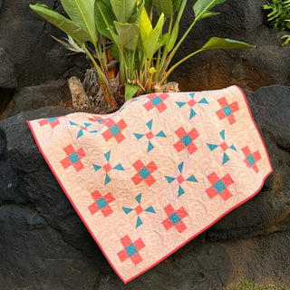 Quilt with coral and teal geometric blocks on a cream background, displayed outdoors on black rocks with green tropical leaves, showing Breezy Shores Quilt Pattern (Download) by Shadow Valley Quilts.