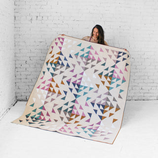 Person holding a colorful Beyond quilt by Eudaimonia Studio which is a geometric-patterned quilt against a white brick wall.