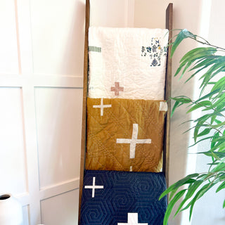 Three quilts with simple cross patterns in cream, mustard, and navy hang on a wooden ladder next to a green plant, showcasing the Callie Quilt Pattern (Download) by Sew & Sew Quilting Co.