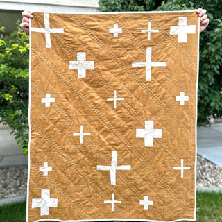 Callie Quilt Pattern by Sew & Sew Quilting Co. baby size: warm rust and white cross pattern quilt held outdoors against a green garden background.