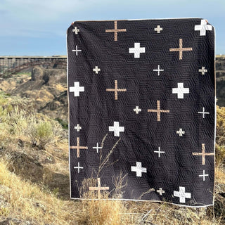 Callie Quilt Pattern by Sew & Sew Quilting Co. cover: black quilt with white and tan cross patterns displayed outdoors against a rocky hillside landscape.