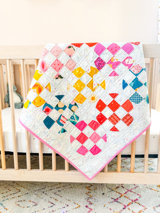 Carlita Quilt Pattern (Download) by Sewspicious displayed as a colorful crib quilt draped over a natural wood crib, featuring bright geometric quilt blocks on a white background.
