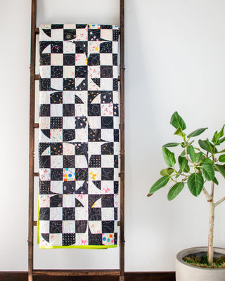 A black and white patchwork quilt with colorful accents hanging on a wooden ladder next to a potted green plant, showcasing the Carlita Quilt Pattern (Download) by Sewspicious.