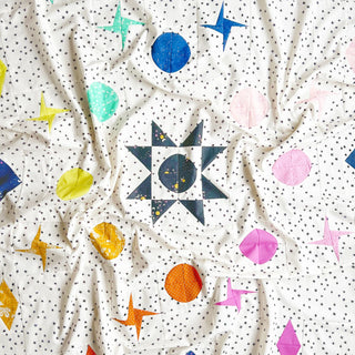 Close-up of fabric featuring star and circle shapes in bright colors on a white background with small black stars, showcasing the Celestial Quilt Pattern (Download) by Lou Orth Designs.