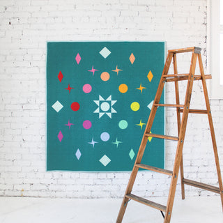 Teal quilt with colorful geometric and star shapes arranged in a circular pattern, displayed on a white brick wall next to a wooden ladder, showcasing Lou Orth Designs Celestial Quilt Pattern (Download).