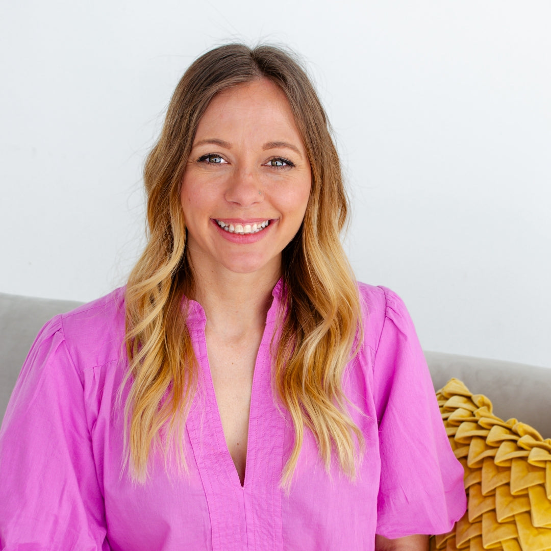 Christina West from Kindred Quilt Co wearing a pink shirt sitting on a couch with a yellow pillow.