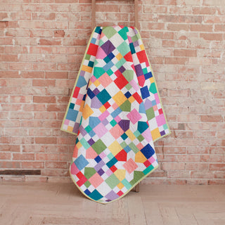 Bright, colorful quilt with geometric patchwork hanging on a ladder against a brick wall, showcasing the Confetti Garden Quilt Pattern (Download) by Straight Stitching.