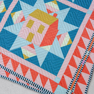 Close-up of a colorful quilt featuring geometric shapes and a house motif in pink, blue, yellow, and coral, highlighting texture and pattern from Toad & Sew’s Cottagecore Quilt Pattern (Download).